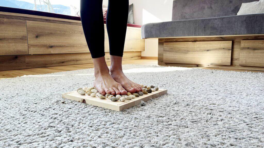 Person steht Barfuß im Wohnzimmer auf dem Fußmassage Brett Big Foot mit Natursteinen und Zirbenholz von Full Balance